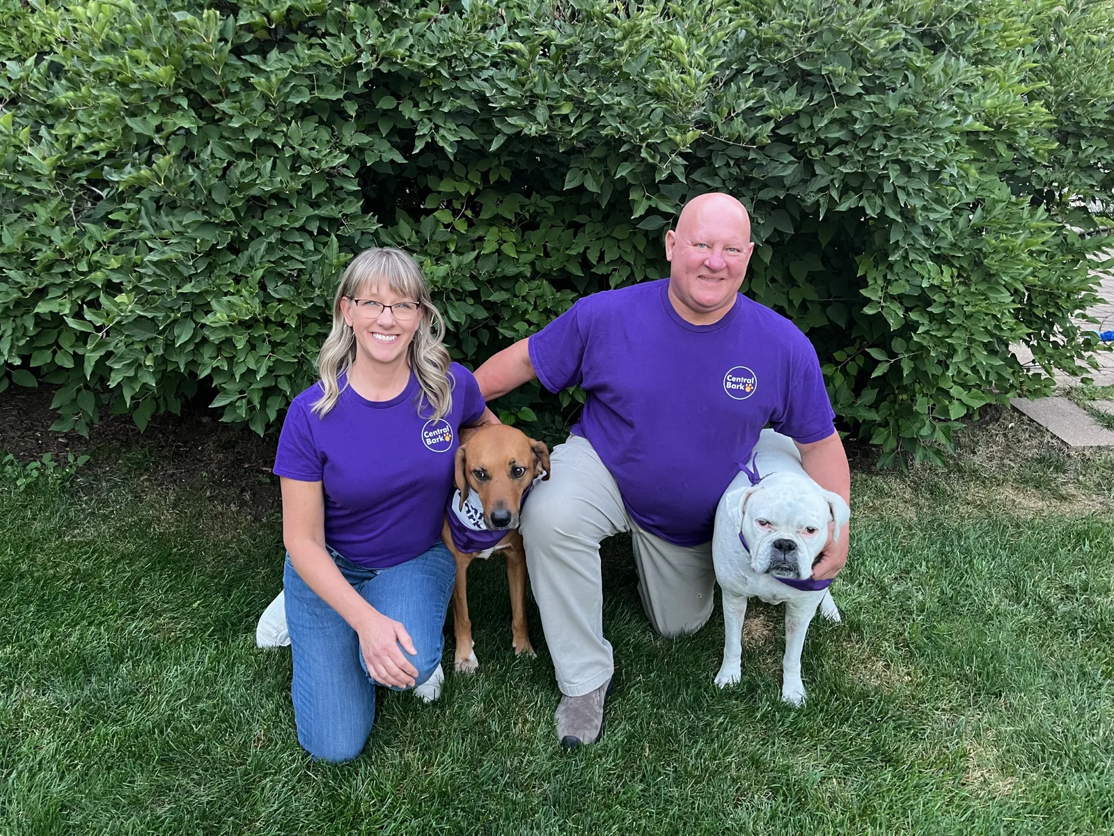 Mike and Beth Carter, owners of Central Bark Aurora Fox Valley, kneeling on the grass with their dogs Lily and Ellie